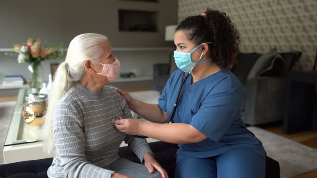 Nurse listenig to senior patient's heartbeat during home visit - wearing protective face mask