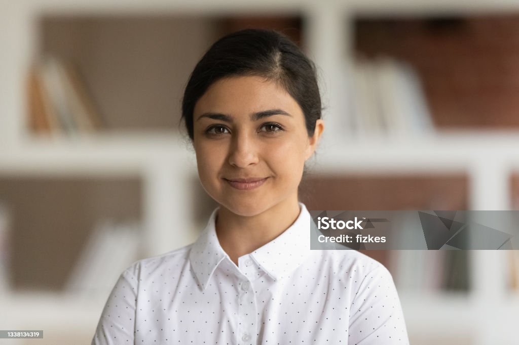 Portrait of young Indian businesswoman or school teacher pose indoors University or school teacher, business lady had shot portrait concept. Young attractive Indian ethnicity woman in white shirt pose in library room with bookshelves on background smile look at camera Headshot Stock Photo