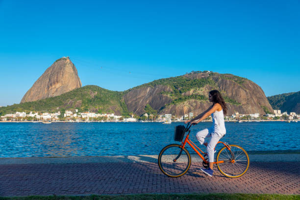 Young woman rides a bicycle in front of Sugarloaf Mountain 24 year old caucasian woman rides on the bike path in front of the Sugarloaf in Flamengo Park, at the border of Guanabara Bay guanabara bay stock pictures, royalty-free photos & images