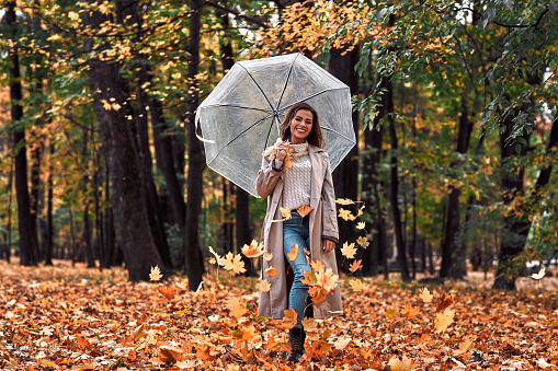 Fun in the autumn park. A young beautiful woman is holding a transparent umbrella from which maple leaves are falling and smiling while looking at the camera.