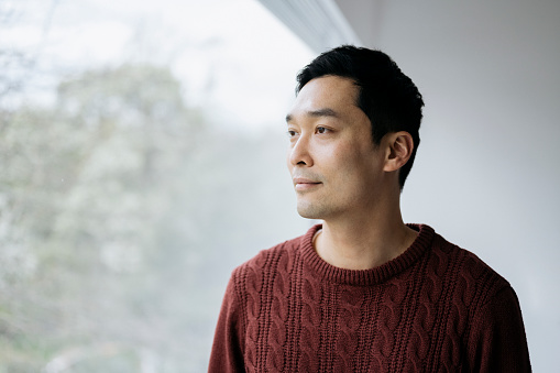 Man with short black hair in rust colored cable stitch sweater standing at picture window and looking away from camera with contented expression.
