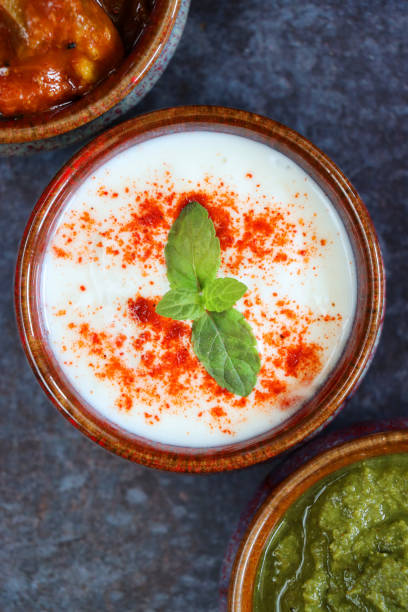 Close-up image of three bowls in a diagonal line containing dips of raita (yoghurt based sauce), mint coriander chutney and green mango pickle, on mottled grey background, Indian cuisine, elevated view