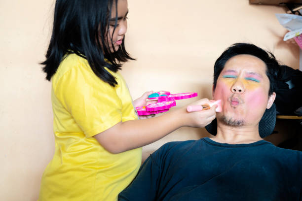 Funny time! Father and his child are playing at home. Cute girl is doing makeup to her dad Funny time! Father and his child are playing at home. Cute girl is doing makeup to her dad, sitting on the bed in the bedroom. Family holiday and togetherness. crazy makeup stock pictures, royalty-free photos & images