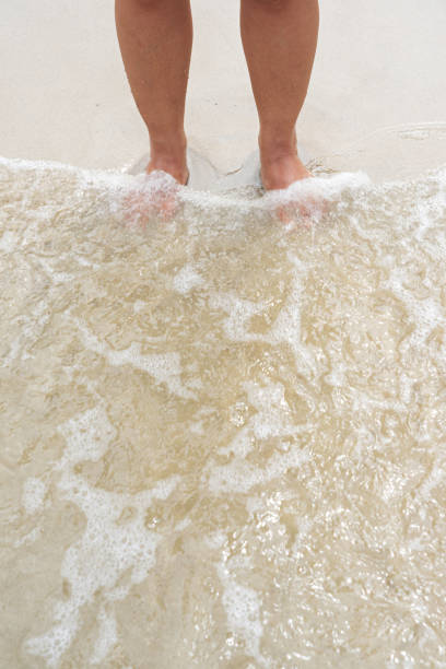 Asian female legs standing on sand beach with small waves touching her feet.