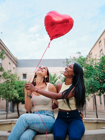 Two women smiling and holding a red heart-shaped balloon while sitting outdoors. Love concept.