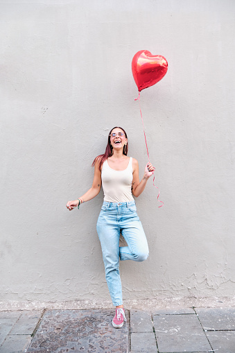 Happy young woman holding a red heart-shaped balloon while leaning on a wall in the street. Love concept.