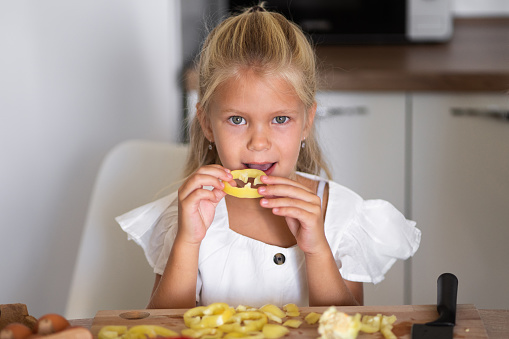 A little blond girl with blue eyes is posing in the kitchen with a slice of yellow bell pepper