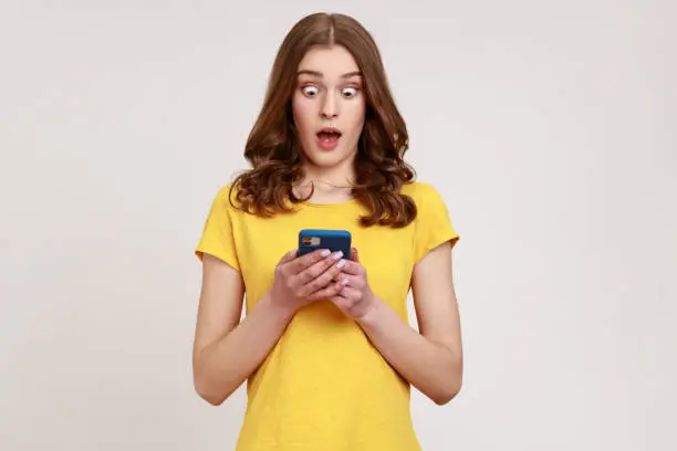 Photo of Mobile connection. Portrait of shocked astonished teenager girl in yellow T- shirt reading shocking post on social network using cell phone.