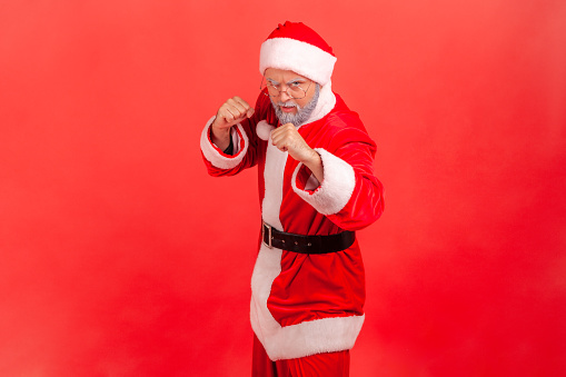 Strict elderly man with gray beard wearing santa claus costume standing with fists and being ready to fight, looking at camera with angry expression.