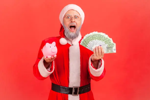 Photo of Excited shocked elderly man with gray beard wearing santa claus costume standing with euro banknotes and piggy bank, big amount of money.