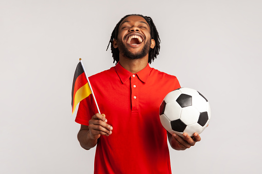 Screaming man wearing red casual T-shirt, football fan sincerely rejoicing, supporting favorite team holding ball and german flag, watching football. Indoor studio shot isolated on gray background.