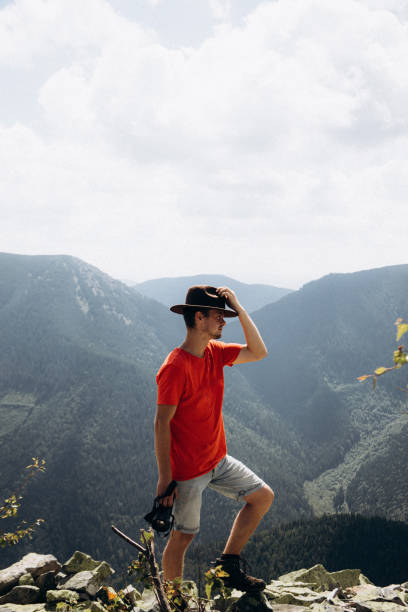 An elderly man, 50-55 years old, adjusts a white hat against a backdrop of mountains and water.