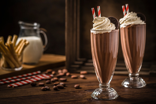 Front view of two chocolate milkshakes with whipped cream and a chocolate cookie on a rustic wooden table