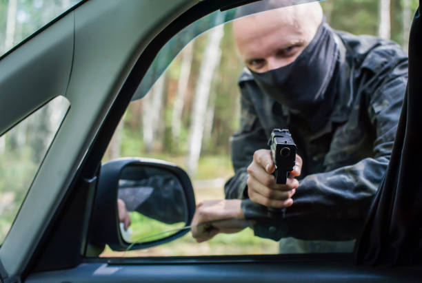 un uomo in mimetica e una maschera da una pistola mira al conducente attraverso il finestrino dell'auto. - thief crime gun hostage foto e immagini stock