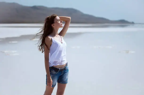 Photo of Young slender beautiful girl in shorts and a T-shirt stands on the shore of a dry salt lake in summer