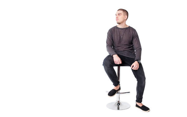 A young attractive guy in gray and black clothes sits on a bar stool on a white background and looks to the side. Copy space. Isolated