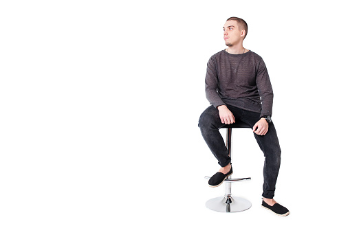 A young attractive guy in gray and black clothes sits on a bar stool on a white background and looks away. Copy space.