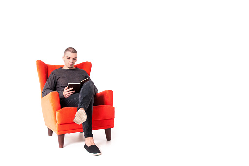 A young attractive guy is sitting on a red armchair and reading a book. Isolated on a white background. Copy space.