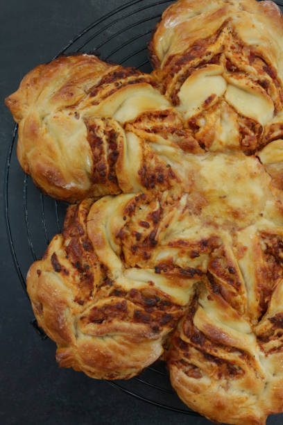 Stock photo showing an close-up view of a golden, freshly, baked mozzarella cheese and tomato puff pastry star, on a black, metal wire cooling rack, against a black background. imagen de la estrella de la pizza de hojaldre recién horneada, dorada, en una rejilla de enfriamiento de alambre de metal negro, circular, relleno de queso mozzarella y salsa de tomate, glaseado con lavado de huevo, fondo negro, vista elevada