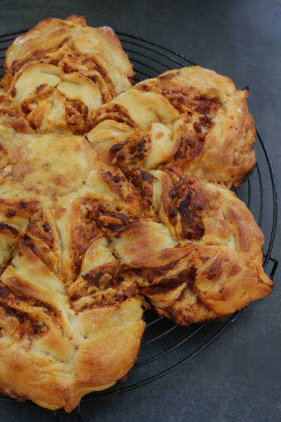 Stock photo showing an close-up view of a golden, freshly, baked mozzarella cheese and tomato puff pastry star, on a black, metal wire cooling rack, against a black background. imagen de la estrella de la pizza de hojaldre recién horneada, dorada, en una rejilla de enfriamiento de alambre de metal negro, circular, relleno de queso mozzarella y salsa de tomate, glaseado con lavado de huevo, fondo negro, vista elevada