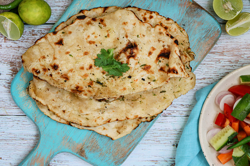 Stock photo showing an elevated view of turquoise, wooden cutting board containing Indian garlic naan flatbread brushed with melted butter (ghee), garnished with coriander leaves, besides blue muslin cloth and plate of chopped vegetables.