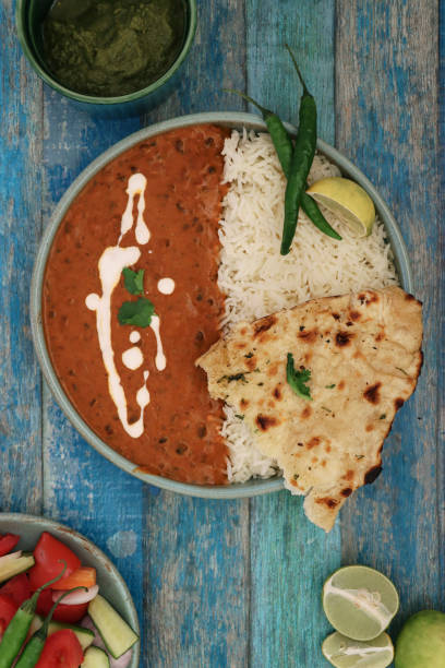 Image of blue bowl containing homemade Dal makhani (black lentils and red kidney bean curry) meal, served with white rice and naan flatbread, lemon slice, green chilli peppers, side salad, mint coriander chutney dip, blue wood grain background, elevated stock photo