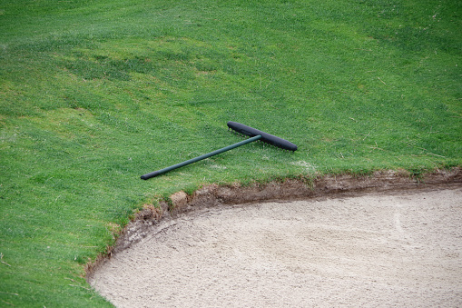 Partial view of a golf course sand trap bunker and a rake next to it on the grass