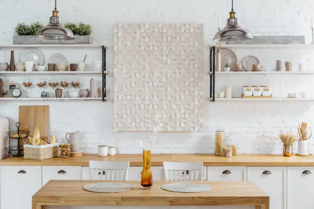 Kitchen interior of white home with brown wood decor. Modern rural house design, wooden furniture with decoration, kitchenware. Cozy scandi apartment style with plant on dining table.