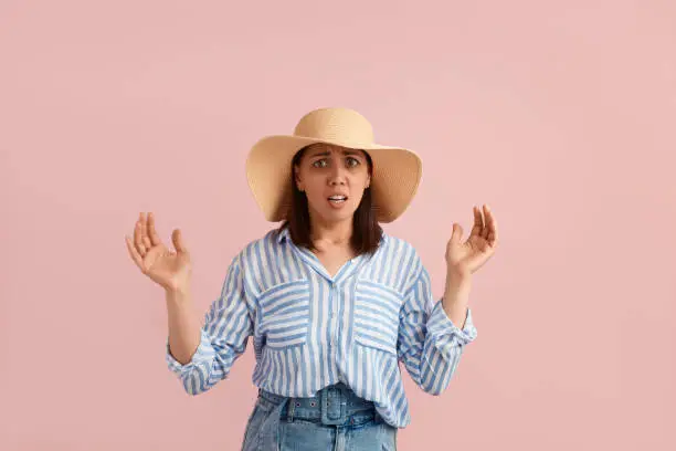 Scared nervous woman with dark hair is afraid of something, keeps mouth open and palms raised, looks at camera, wears straw hat, striped shirt, jeans, on pink background. Summer emotions concept
