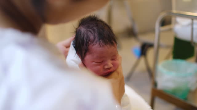 A woman of asian ethnicity in the hospital holding her new born baby boy. Here she is reclining and holding the child close to her chest comforting him.