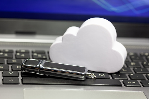 Cloud data storage technology, storing data in the cloud abstract. Cloud and a usb memory stick, pen drive laying on a laptop keyboard. Online private personal data backup service tech simple concept