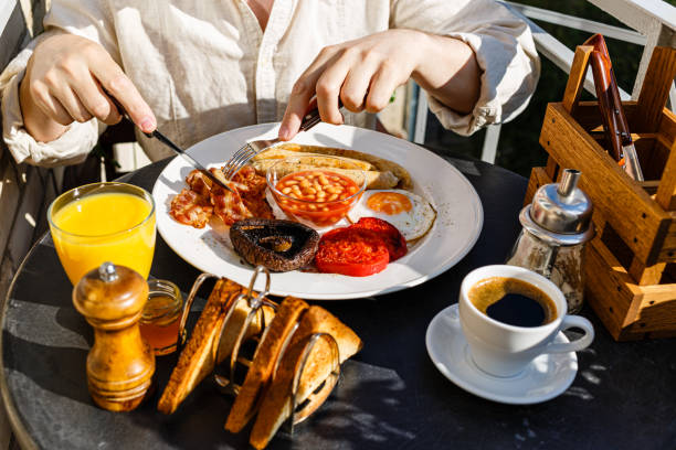 mężczyzna jedzący pełne podsmaż angielskie śniadanie z jajkami sadzonymi, kiełbaskami, grzybami portobello, fasolą, tostami i kawą podawane na okrągłym stole w słoneczny letni poranek - eggs fried egg frying pan fried zdjęcia i obrazy z banku zdjęć