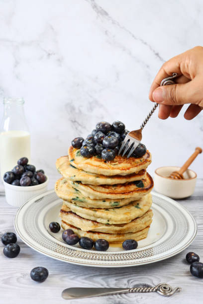 istock Image of unrecognisable person using metal fork to cut into stack of fluffy, American-style blueberry pancakes, pancakes decorated with fresh blueberries drizzled with dripping maple syrup on white plate, marble effect background, healthy eating 1338305423