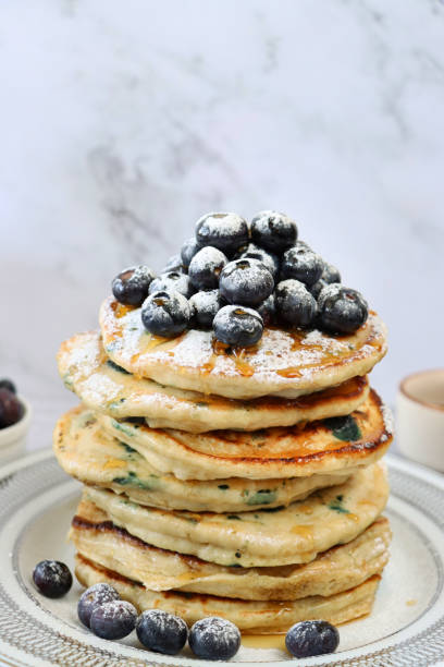 istock Image of fresh blueberries drizzled with dripping maple syrup and dusted with sieved icing sugar on top of stack of fluffy, American-style blueberry pancakes on a white plate, marble effect background, healthy eating 1338306050