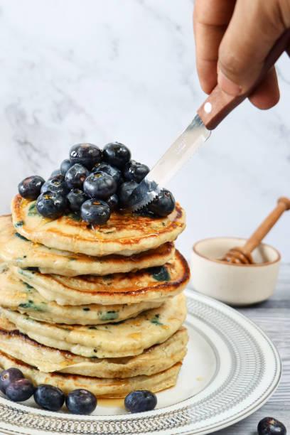 istock Image of unrecognisable person using serrated knife to cut into stack of fluffy, American-style blueberry pancakes, pancakes decorated with fresh blueberries drizzled with dripping maple syrup on white plate, marble effect background, healthy eating 1338306659
