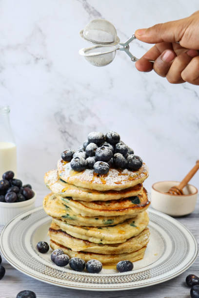 Image of unrecognisable person using sieve to sift icing sugar over stack of fluffy, American-style blueberry pancakes, pancakes decorated with fresh blueberries drizzled with dripping maple syrup on white plate, marble effect background, healthy eating
