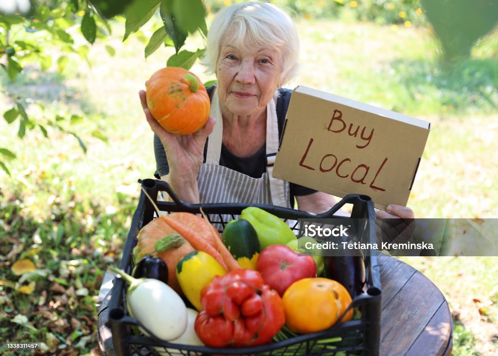 An elderly gray-haired woman sells fresh seasonal vegetables at a local farmers' market. Buy local agricultural products An elderly gray-haired woman sells fresh seasonal vegetables at a local farmers' market. Local business support. Buy local non-GMO agricultural products. Farmer's Market Stock Photo