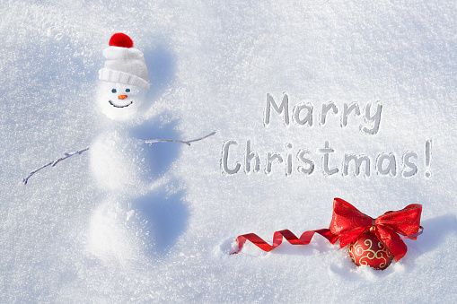 Snowy winter card wallpaper for Christmas. Funny snowman of snowdrifts on snow and a red ball with a bow outdoors on nature in sunny day and inscription Merry Christmas., flat lay.