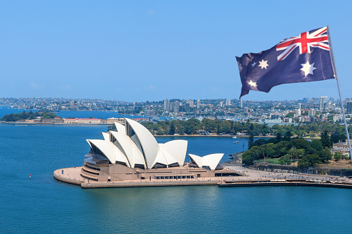 Sydney, New South Wales, Australia - September 4th, 2021:  An Australian flag blows in the wind from an elevated view of the Opera House. The Sydney Opera house is considered one of the major landmarks of Australia and a big tourist attraction.