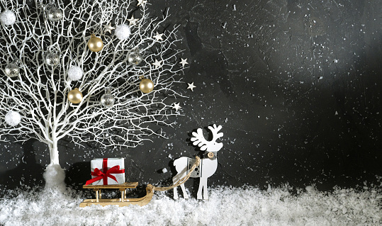 Festive Christmas background template - \nbright black scene decoration color with white tree in snow and toy reindeer with wooden sleigh with box gift with red ribbon, copy space, top view.