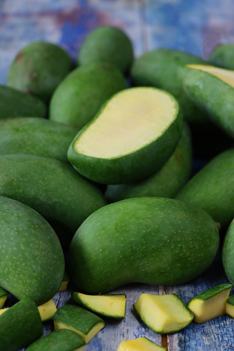 Stock photo showing close-up, elevated view of pile of fresh, whole, tropical raw mango fruit with one sliced in half, against a blue wood background.