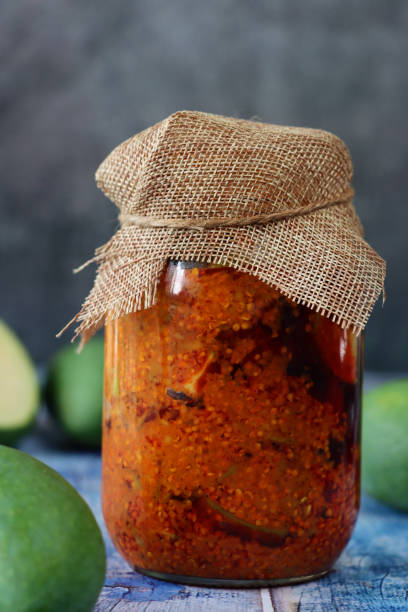 Image glass jar of green mango chutney recipe with hessian lid, pickle with chopped raw mango, seeds, spices and mineral condiments, blue wood grain surface, grey background, focus on foreground Stock photo showing close-up view of green (raw) mango chutney recipe in a glass jar covered with hessian on blue wood grain surface. Ingredients include, powdered turmeric, nigella onion seeds, red chilli powder, fenugreek seeds, pickling salt and mustard seeds. indian pickles in jar stock pictures, royalty-free photos & images