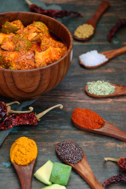 Stock photo showing a close-up view of wooden spoonfuls of condiments for green mango chutney recipe. Ingredients include, powdered turmeric, nigella onion seeds, red chilli powder, fenugreek seeks, pickling salt and mustard seeds. imagen de un tazón de madera que contiene chutney de mango verde (crudo), rodeado de cucharas de madera de especias y minerales utilizados para dar sabor al pepinillo, chiles rojos secos, condimentos, ingredientes, muselina gris / verde, vista elevada, en