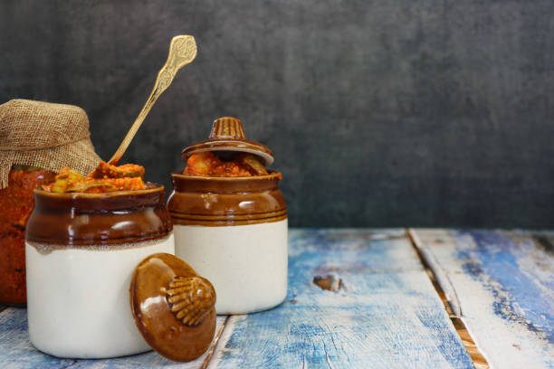 Image ceramic pickle lidded pots, hessian covered glass jar of green mango chutney recipe, metal spoon, pickle with chopped raw mango, seeds, spices and mineral condiments, blue wood grain surface, grey background, focus on foreground, copy space stock photo