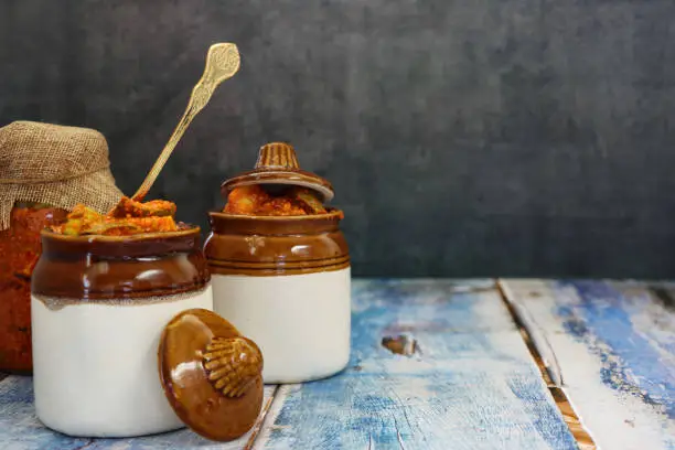 Stock photo showing close-up view of green (raw) mango chutney recipe in glass jar covered with hessian and lidded, ceramic pickle pots on blue wood grain surface. Ingredients include, powdered turmeric, nigella onion seeds, red chilli powder, fenugreek seeds, pickling salt and mustard seeds.