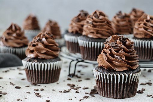 Stock photo showing a close-up view of a batch of freshly baked, homemade, chocolate cupcakes in paper cake cases on a circular, black, metal wire cooling rack. The cup cakes have been decorated with swirls of chocolate piped icing and chocolate pieces.