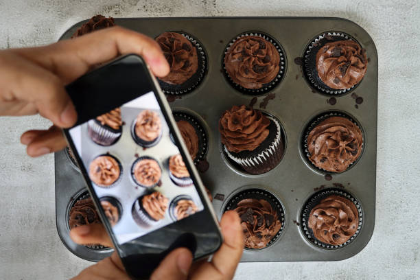 image of unrecognisable person using smartphone  to photograph muffin cake tin containing homemade, chocolate cupcakes in paper cake case, chocolate butter icing piped swirl topped with chocolate pieces, cakes lying on side, elevated view - muffin blueberry muffin blueberry butter imagens e fotografias de stock
