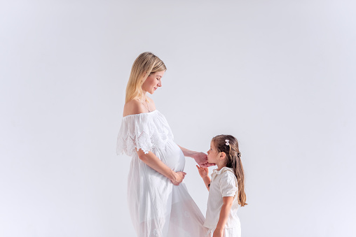Little girl kisses hugs pregnant mother on an isolated white background. The older sister is waiting for the baby. Healthy young woman carries child, hugs her daughter. Happy modern family. Copy space