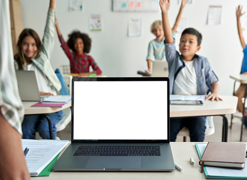 Laptop computer white blank empty mockup screen on teachers table with elementary junior children students raising hands in classroom background. Education software website technology ads concept.