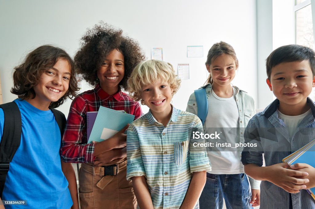 Happy diverse junior school students children looking at camera in classroom. Happy diverse junior school students children group looking at camera standing in classroom. Smiling multiethnic cool kids boys and girls friends posing for group portrait together. Child Stock Photo